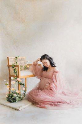 A pregnant mother sitting next to a rattan char with a bouquet of flowers wearing pink