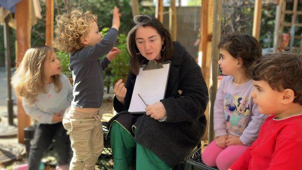 A group of children are discussing facial features to add to their portraits. A child observes Teacher Laila's hair for her portrait.