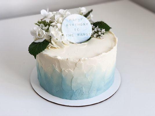 Signature cake with two tone pastel blue textured cream topped with fresh flowers and a customized inscription