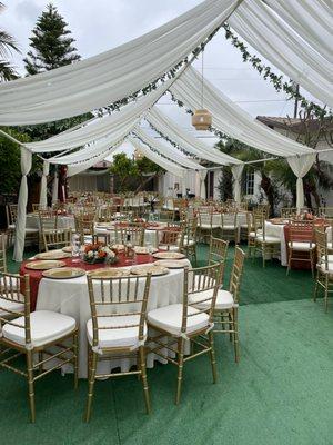 Wedding setup: draped tent, round tables, ivory linen, runner, charger plates, gold chiavari chairs