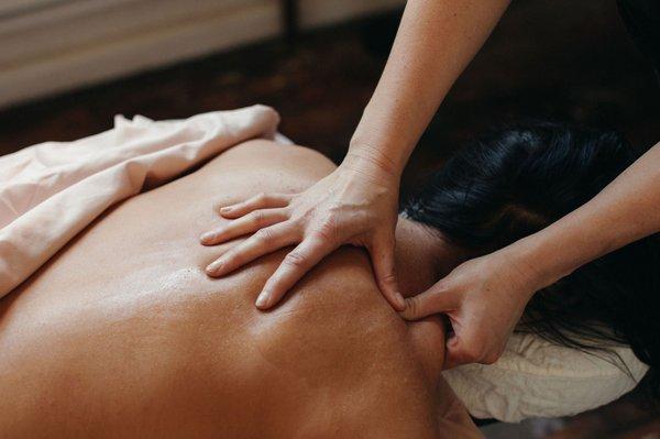 A person lays prone with their back exposed on a massage table while a practitioner's hands work the muscle sin the upper shoulder.