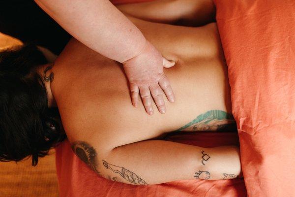 A woman with tattoos lays face down on warm salmon colored sheets while a hand gently touches her back.