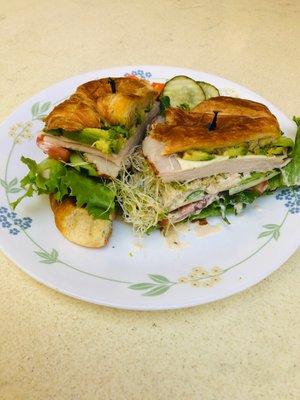 $4.95 (2021 pricing) croissant sandwich generously stuffed with avocado, turkey, alfafa, house dressing, tomatoes, cucumber, lettuce+onions