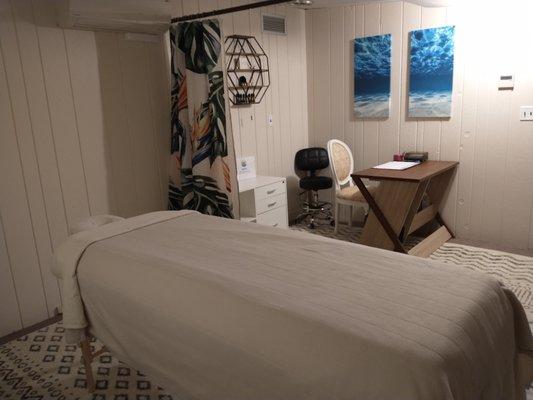 Photo of massage table, open curtain and office space with desk and chair.