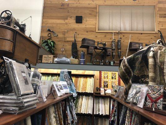 More vintage sewing machines and vintage vacuums above the wall of bolts of fabrics.