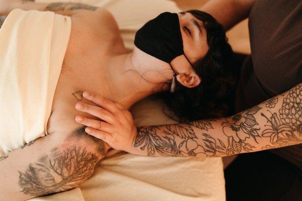 A woman lays supine during a massage, wearing a black face mask, while a practitioner massages her shoulders.