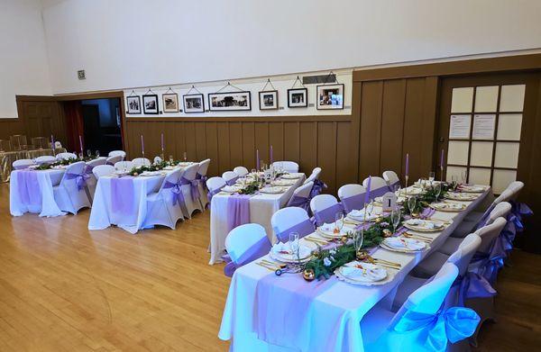 Rectangular tables lined up in a banquet hall. Tables and chairs are decorated in white and purple.