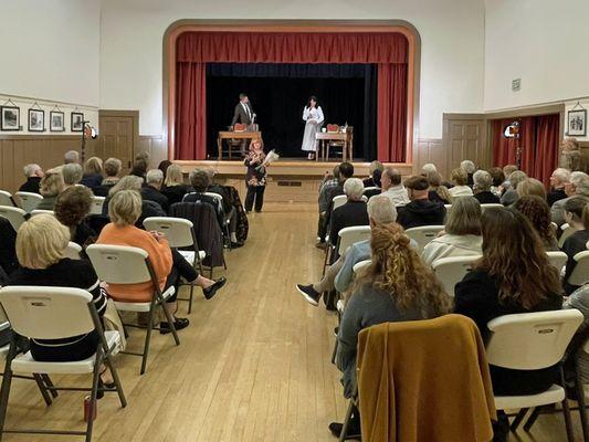 Two people are standing on a stage. A women is standing in front with a mic. An audience is watching