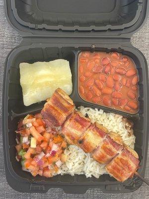 Plate with white rice, brown beans, yuca, chicken bacon wrapped barbecue and vinagrete.