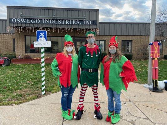 Three staff members dressed as elves pose in front of the Oswego Industries building.